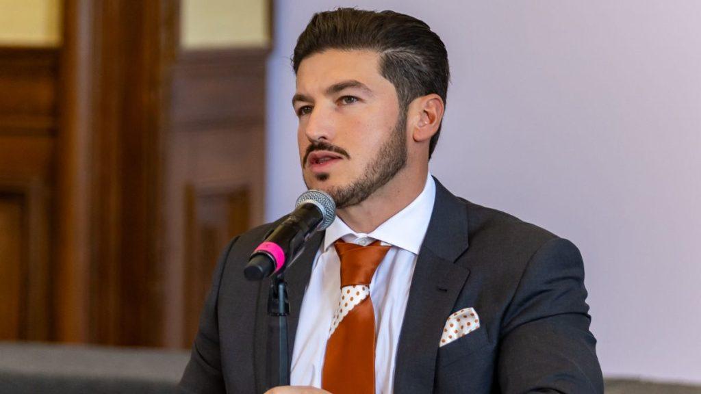 Man in a Dark Suit and Orange Tie Speaks into a Microphone at a Formal Event. - Justicia Regia
