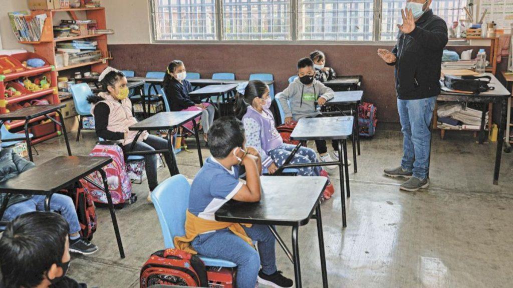a Teacher Wearing a Mask Stands at the Front of a Classroom, Signaling with His Hands As Students at Individual Desks Listen, All Wearing Masks. - Justicia Regia