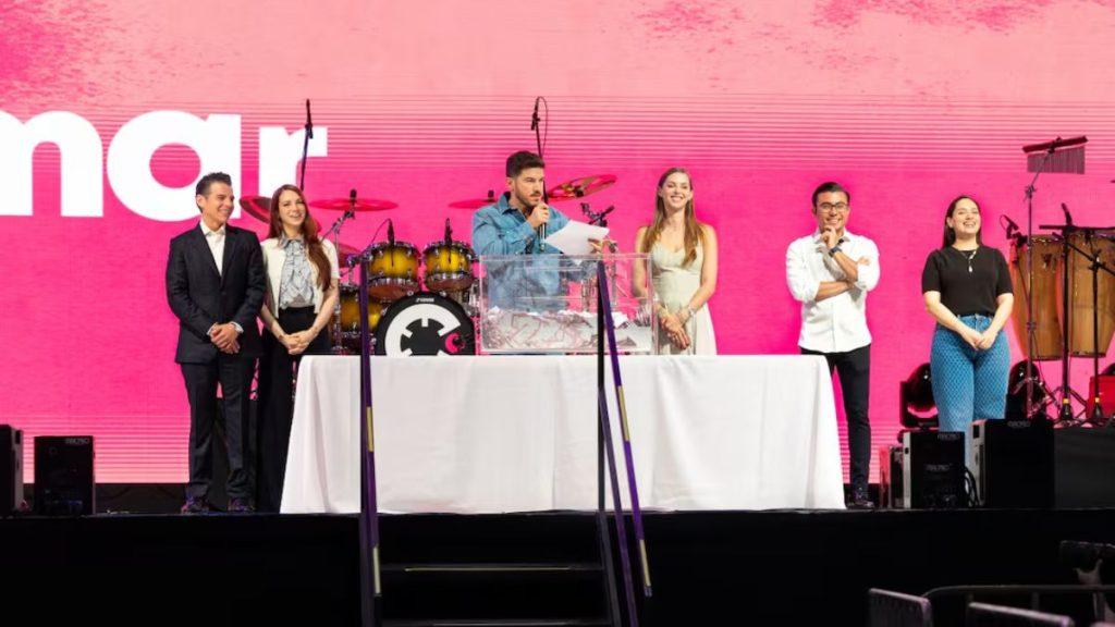 Group of Six People on a Stage Behind a White-clothed Table, Speaking into Microphones at a Pink Backdrop with Drums in the Background. - Justicia Regia
