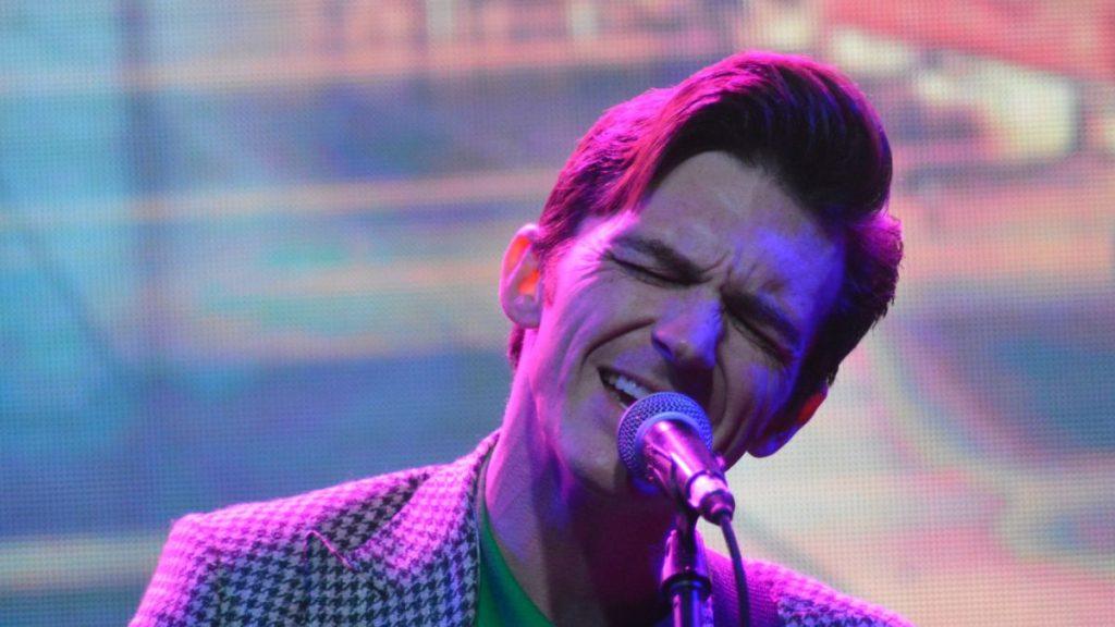Male Singer with a Retro Hairstyle Performing into a Microphone on a Stage, Wearing a Checkered Blazer Under Colorful Lights. - Justicia Regia