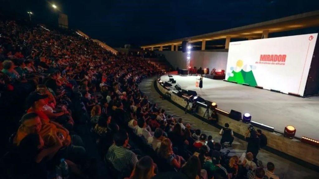 Inauguracion-del-parque-del-agua - Justicia Regia Large Audience in an Open-air Theater Watching a Presenter on Stage with a Bright Screen Displaying Mirador. - Justicia Regia