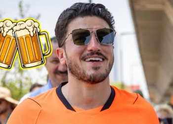 Smiling Man in an Orange Shirt Wearing Sunglasses at an Outdoor Event, with a Cartoon Beer Mugs Sticker on His Left. - Justicia Regia