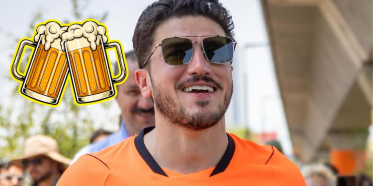 Smiling Man in an Orange Shirt Wearing Sunglasses at an Outdoor Event, with a Cartoon Beer Mugs Sticker on His Left. - Justicia Regia