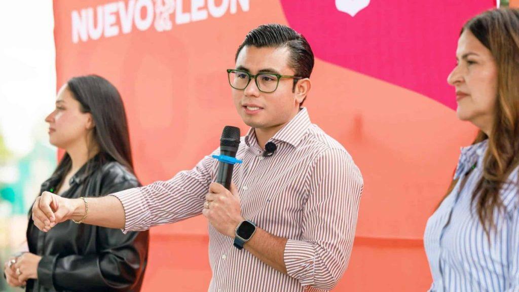 Man in a Striped Shirt and Glasses Speaks into a Microphone at a Panel or Public Talk, with Two Women in the Background and a Pink/orange Backdrop Behind Him. - Justicia Regia