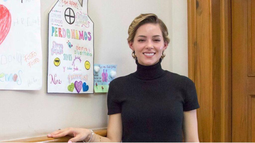 Smiling Woman in a Black Turtleneck Stands Beside Colorful Handmade Posters on a Classroom Wall. - Justicia Regia