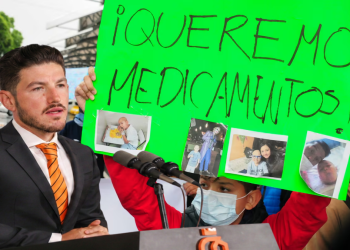 Man in a Suit at a Podium During a Protest; a Participant Holds a Bright Green Sign Reading 'queremos Medicamentos' with Photos Attached. - Justicia Regia