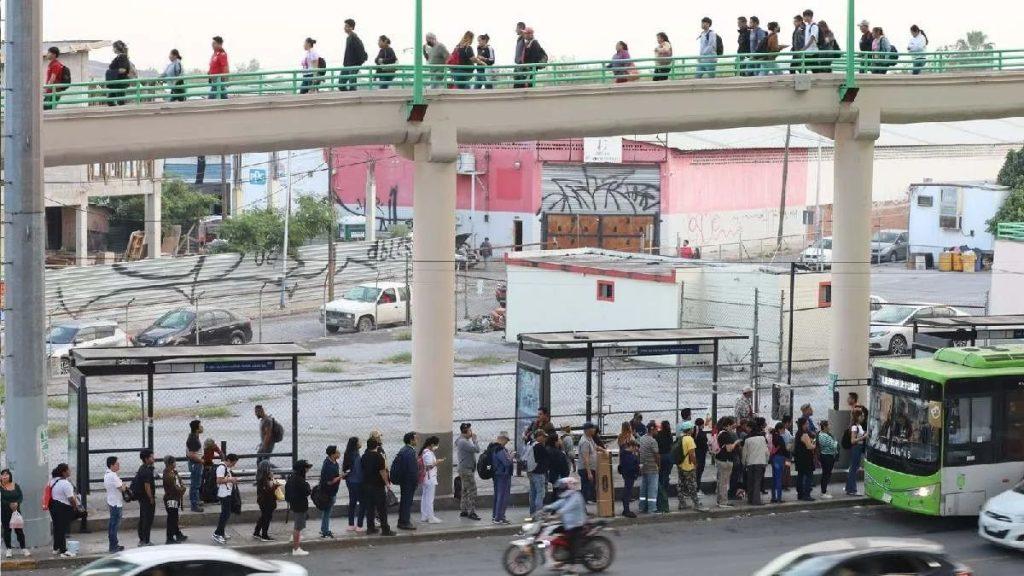 People Stand in a Long Queue at a Bus Stop Under an Elevated Pedestrian Walkway; a Green Bus Sits at the Curb with Cars and Motorbikes in Traffic in a Busy Urban Street. - Justicia Regia