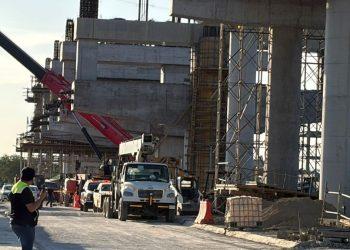 Construction Site with Large Concrete Framework, Scaffolding, and a Red Crane; Trucks and Workers on a Road Beside the Site. - Justicia Regia