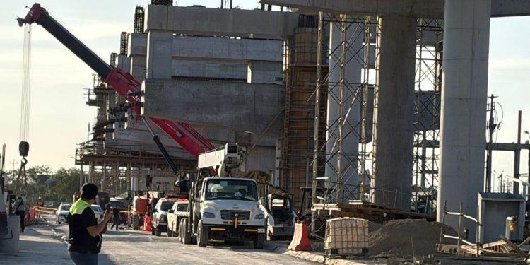 Construction Site with Large Concrete Framework, Scaffolding, and a Red Crane; Trucks and Workers on a Road Beside the Site. - Justicia Regia