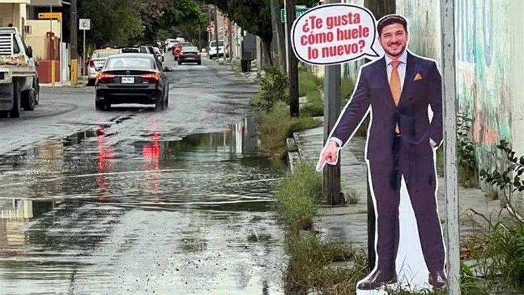 Life-size Cutout of a Man in a Suit with a Speech Bubble Reading '¿te Gusta Cómo Huele Lo Nuevo?' on a City Street with a Puddled Road in the Foreground. - Justicia Regia