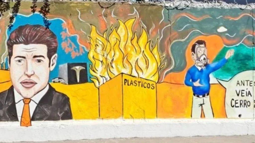 Mural Showing a Man in a Suit on the Left, Burning Crates Labeled Plasticos in the Center, and a Man in Blue Speaking on the Right About Plastics Waste. - Justicia Regia